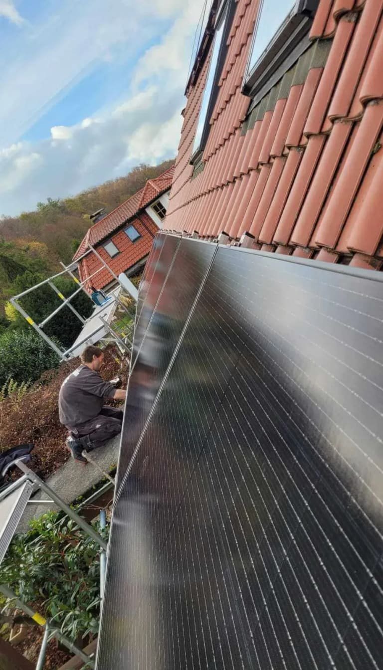 Solarmodule dicht an der Ziegelkante eines Steildachs, dahinter ein Monteur auf dem Gerüst